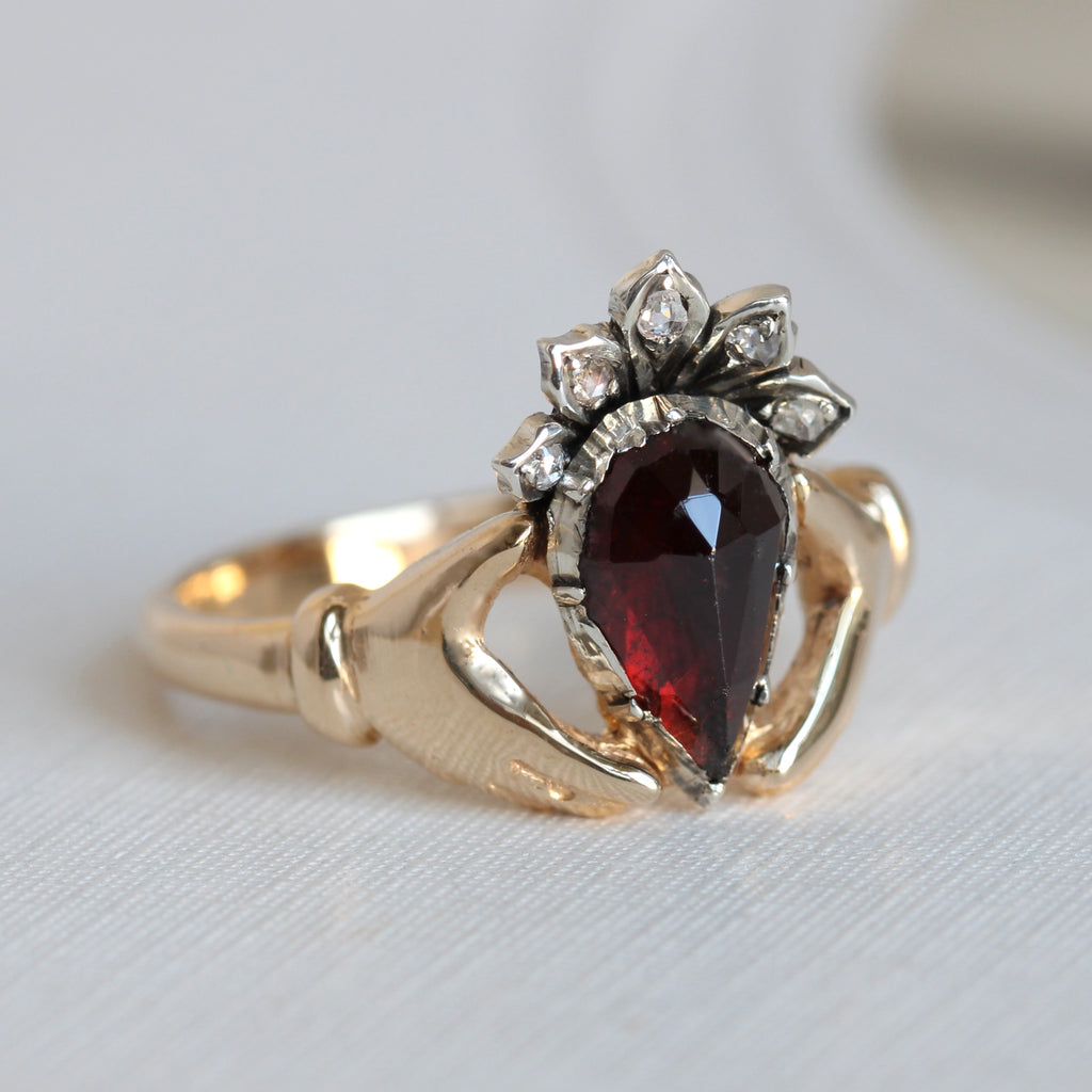 yellow gold ring with hands holding a garnet heart that's set in silver with a silver crown dotted with diamonds 