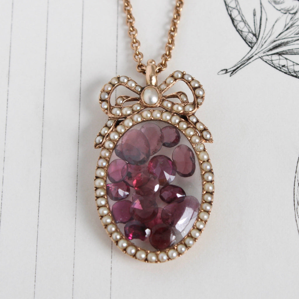 Yellow gold victorian pendant with a pearl bezel and bow design filled with loose faceted garnets behind a glass dome.