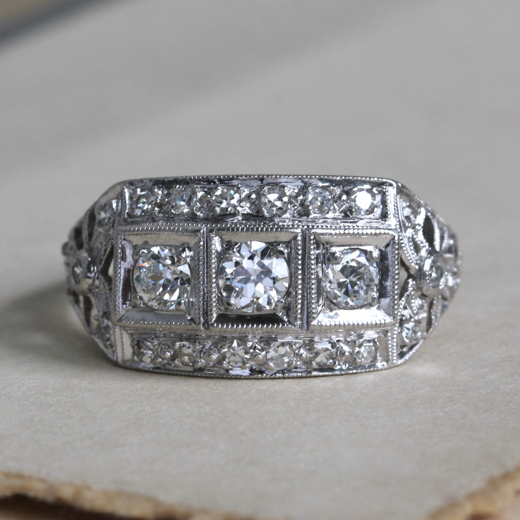 antique platinum ring with three diamonds horizontally across the face and a frame of smaller diamonds, with little filigree bows at each shoulder