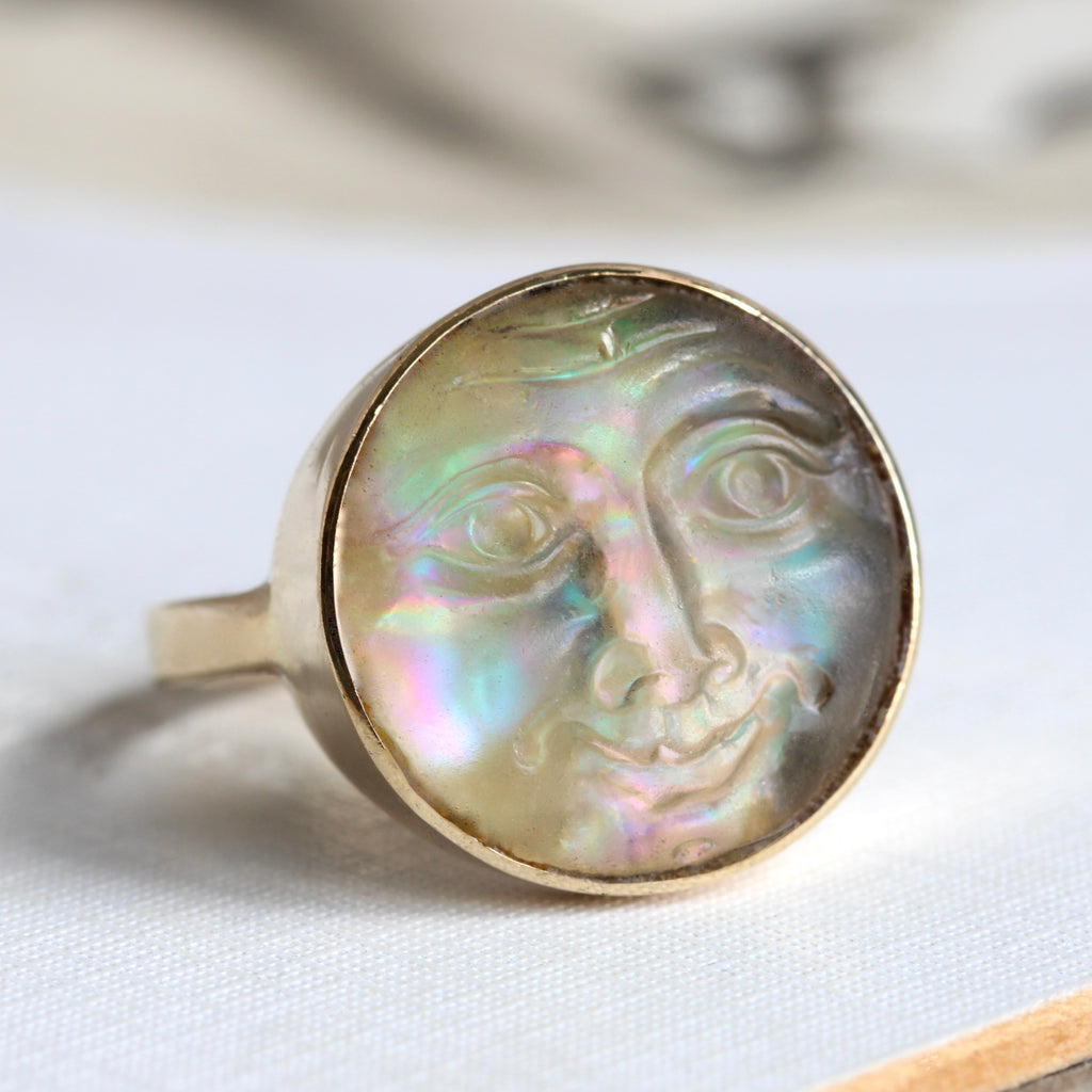 man in the moon face carved in glass that has a shimmering rainbow appearance mounted as a yellow gold ring 