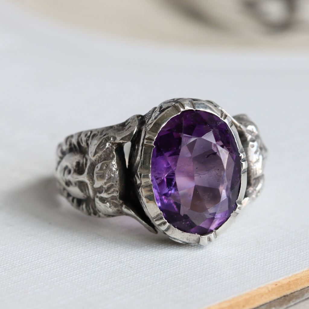 silver ring with a smiling face carved onto the shoulders that wears winged headdress and has entwined snakes for the band, set with an amethyst