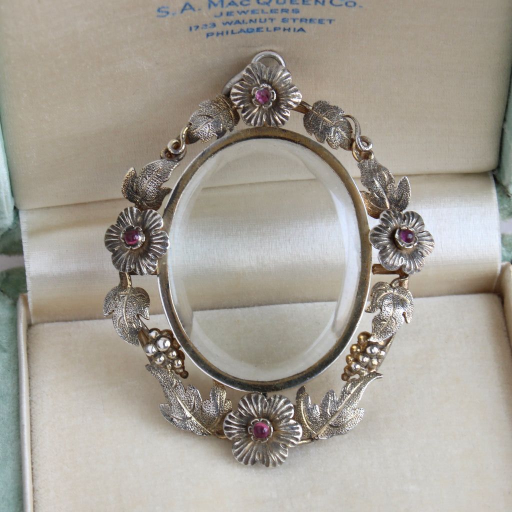 locket in silver with a wreath of flowers, grapes and leaves around a compartment that has glass front and back