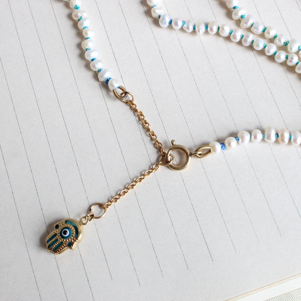 small slightly irregular oval pearls knotted on teal tp blue ombre silk with a gold and blue glass hamsa charm