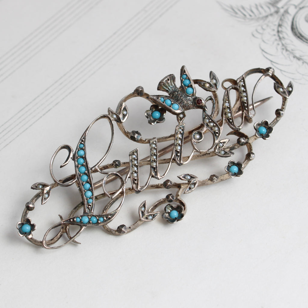 a brooch of the name luisa in fancy script cut out of sterling silver with pearl and turquoise stones set along the lines of the letters