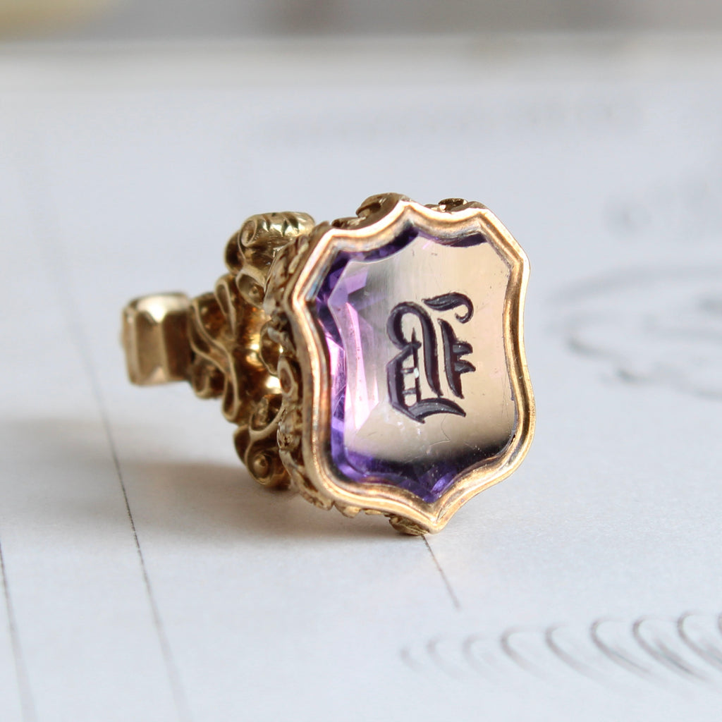 sheild shaped purple stone carved with a letter b in a gothic style font set in a fancy gold fob pendant