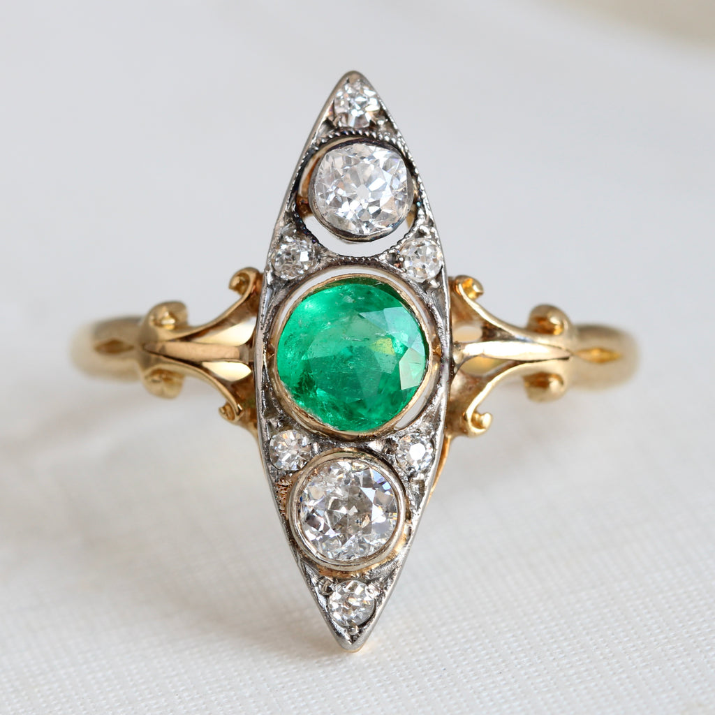 ring with a marquise or navette shaped face set with a round emerald and two large diamonds  north and south, surrounded by a mix of smaller size diamonds with a yellow gold shank