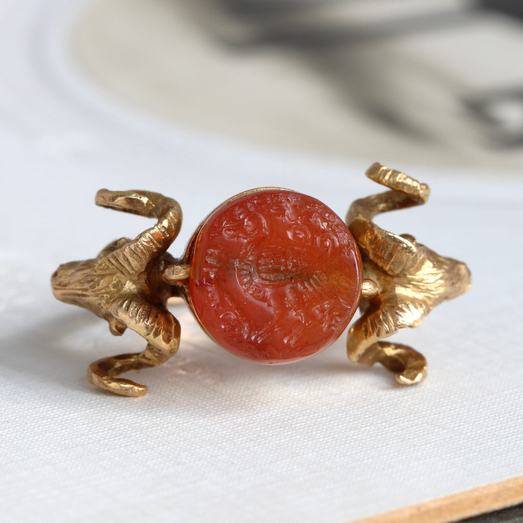 yellow gold ring with a rams head on each shoulder set with an ancient carnelian stone carved with a bird