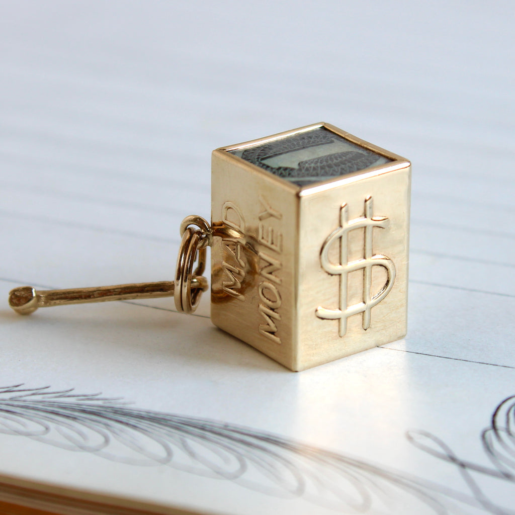 gold charm that looks like a box with a dollar bill inside and a tiny hammer