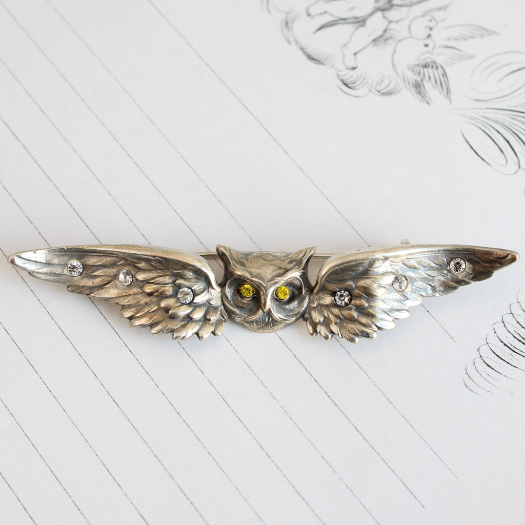 silver owl brooch with an owl head and outstretched wings on either side, accented by white and yellow rhinestones