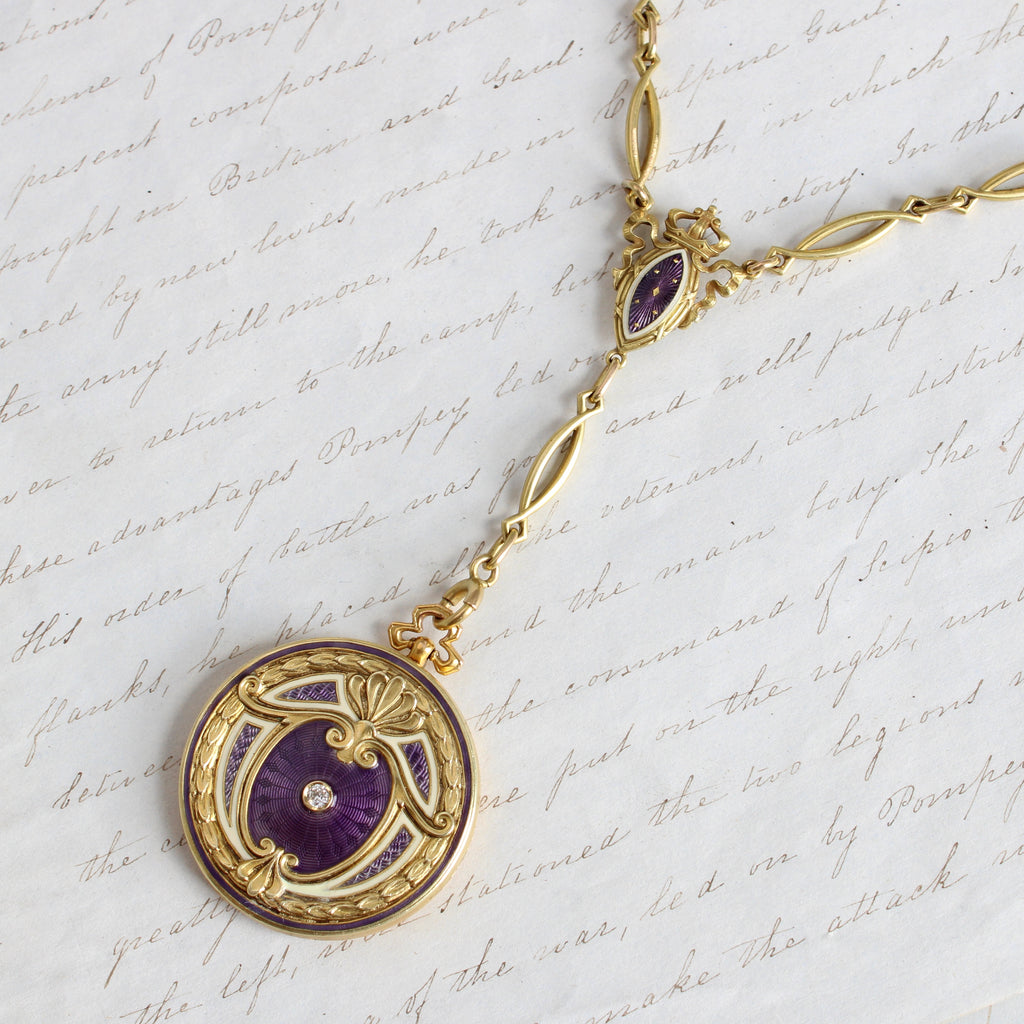 14k yellow gold locket necklace circa 1910; the chain has curved marquise-shaped links alternating with stations enameled in velvety purple and creamy white. The locket has an acanthus leaf border and scrolled center elements on the cover, framing a matching purple guilloché enamel field, and old cut diamond accent