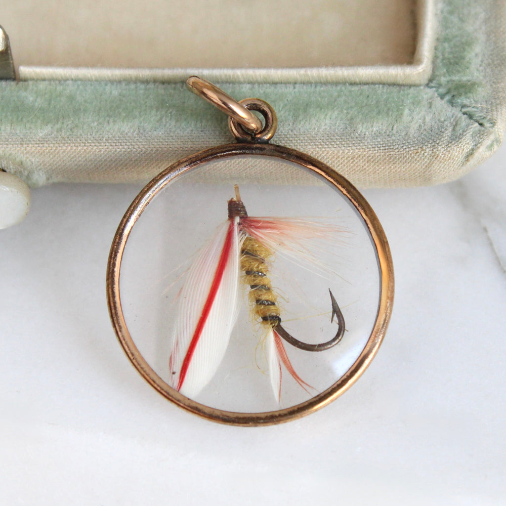 fishing lure made of feathers suspended  inside a glass paned circle locket with rose gold colored metal frame