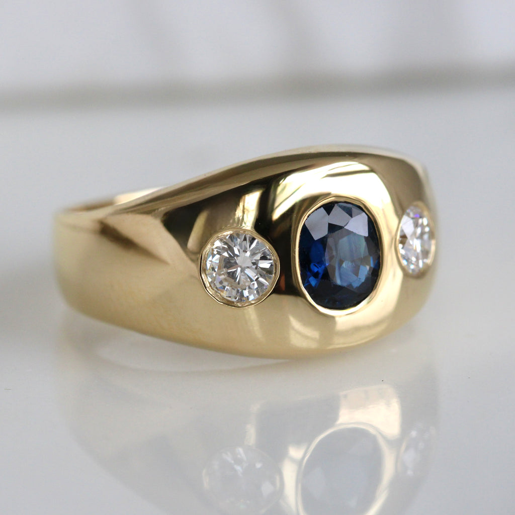 A vintage yellow gold flush mount ring with a blue oval sapphire centerpiece and two twin brilliant-cut diamonds on the sides.
