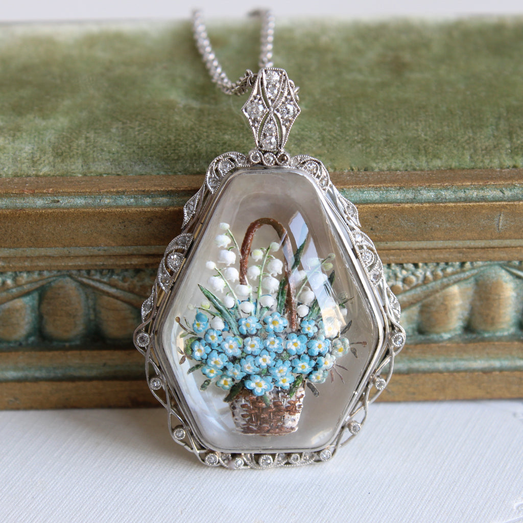 antique pendant with the image of a basket filled with blue forget me nots and white lily of the valley, carved into the back of a crystal and painted, in a platinum filigree fgrame studded with diamonds