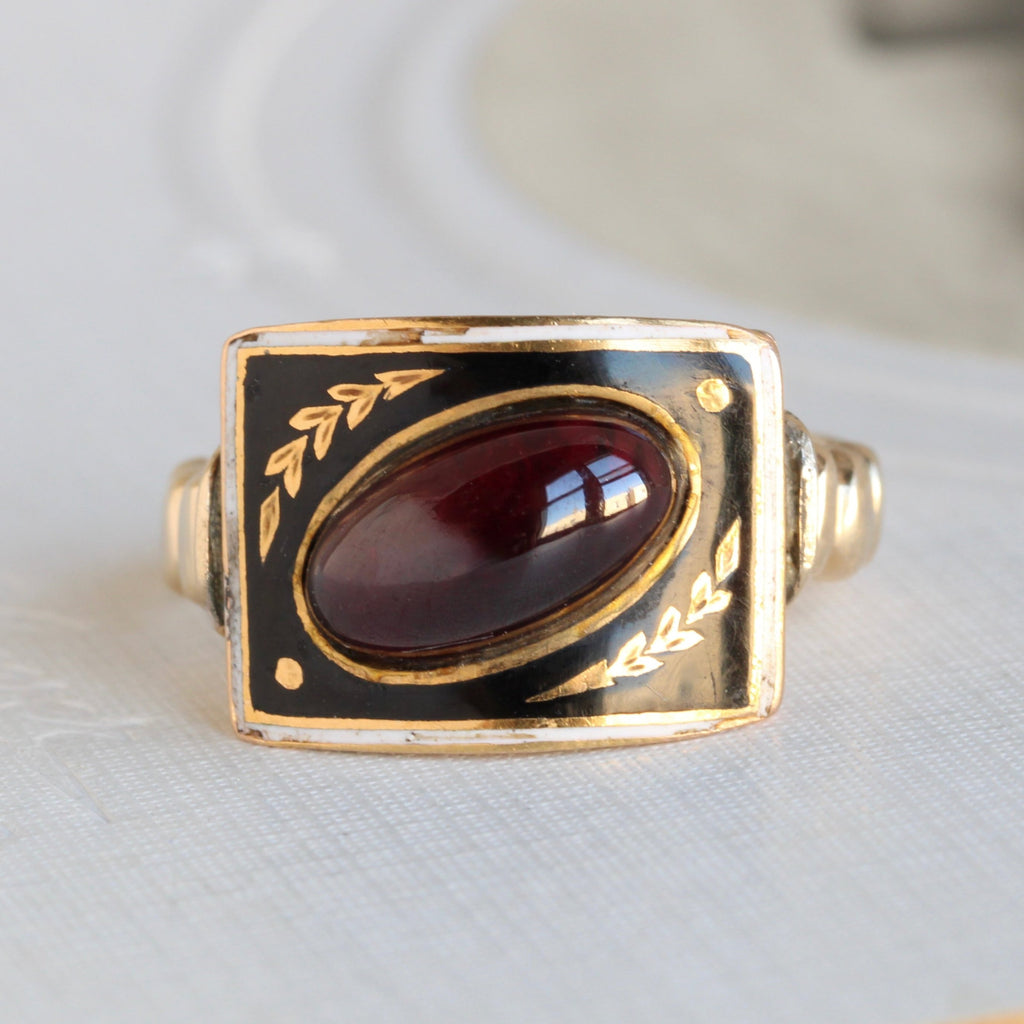 gold ring with a rectabgular face enameled in black with a white border and an oval garnet cabochon set diagonally across the face.