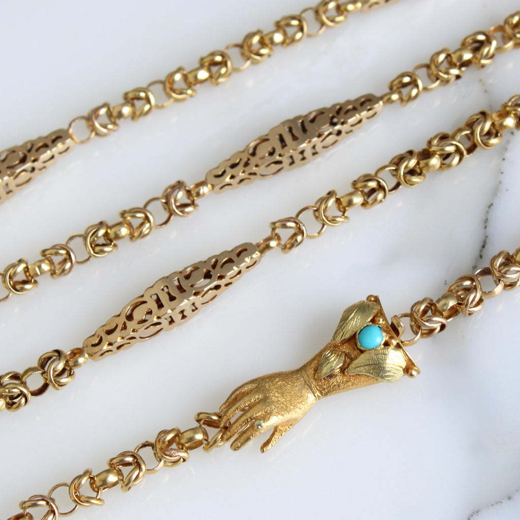 long gold chain with long thin barrel links alternating with cable chain and a gold claps shaped like a hand.