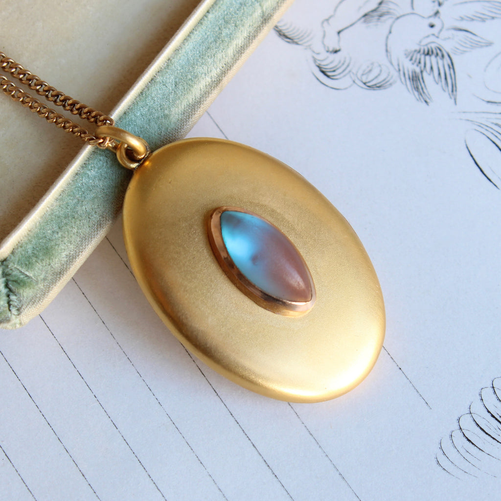 yellow gold metal oval locket set with a blue to pink glass marquise shaped cabochon on a gold chain