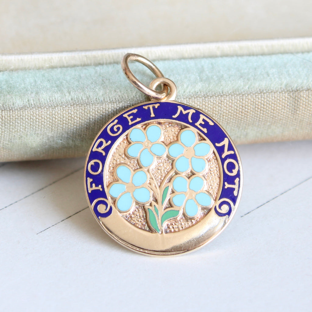 round yellow gold charm enameleed in baby flowers and the words forget me not in gold against a dark blue backround