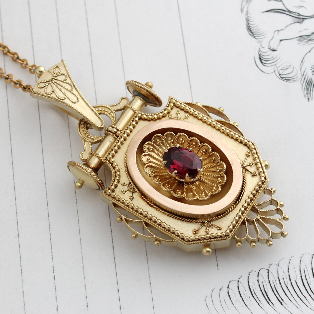 antique yellow gold locket with intricate applied filigree decoration, a large decoarted bail, and a garnet in the center of the front cover, opens in the back to a compartment. On a gold chain