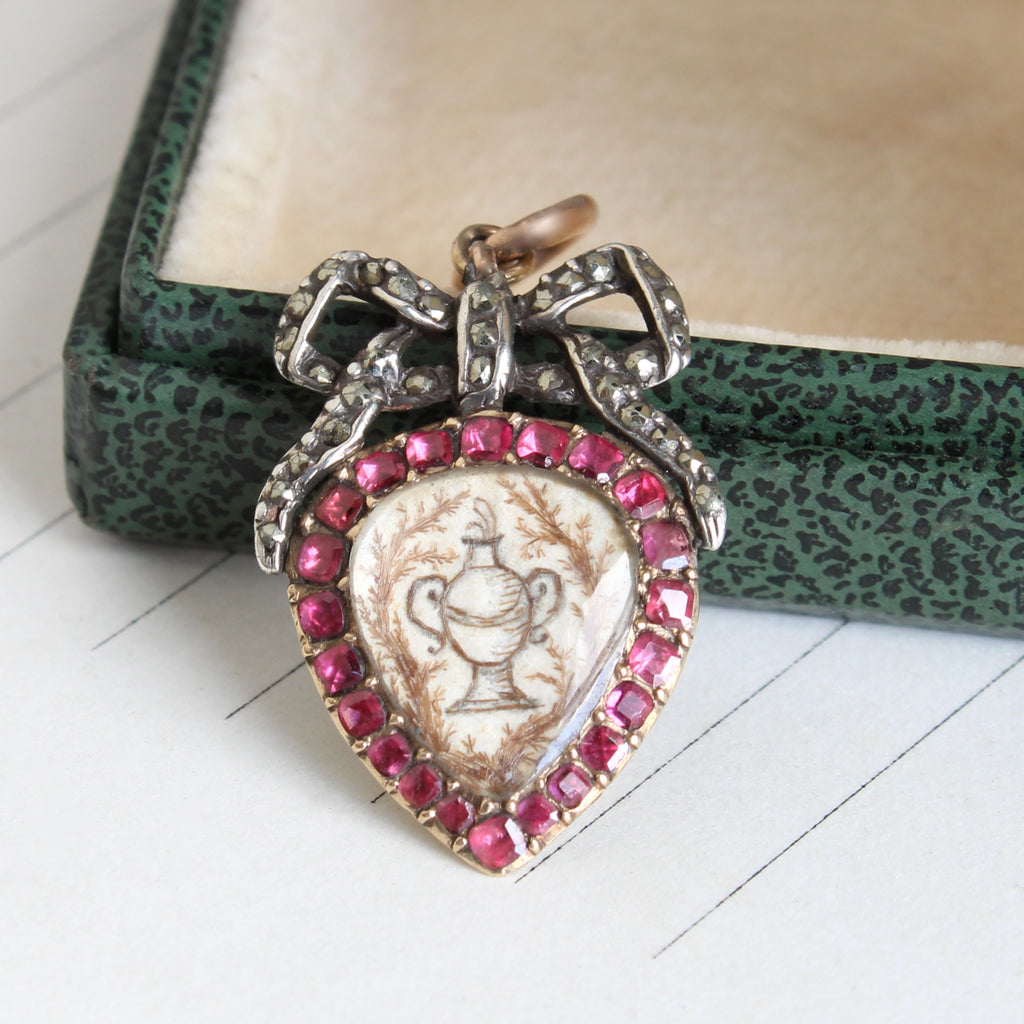 small locket with a bow atop a heart shaped compartment holding a miniature painting of a funeral urn, set with garnets and marcasites 