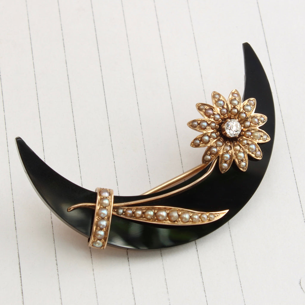 a brooch with a black carved stone crescent moon that has a gold daisy attached across the curve, the daisy set with pearls and a diamond in the center