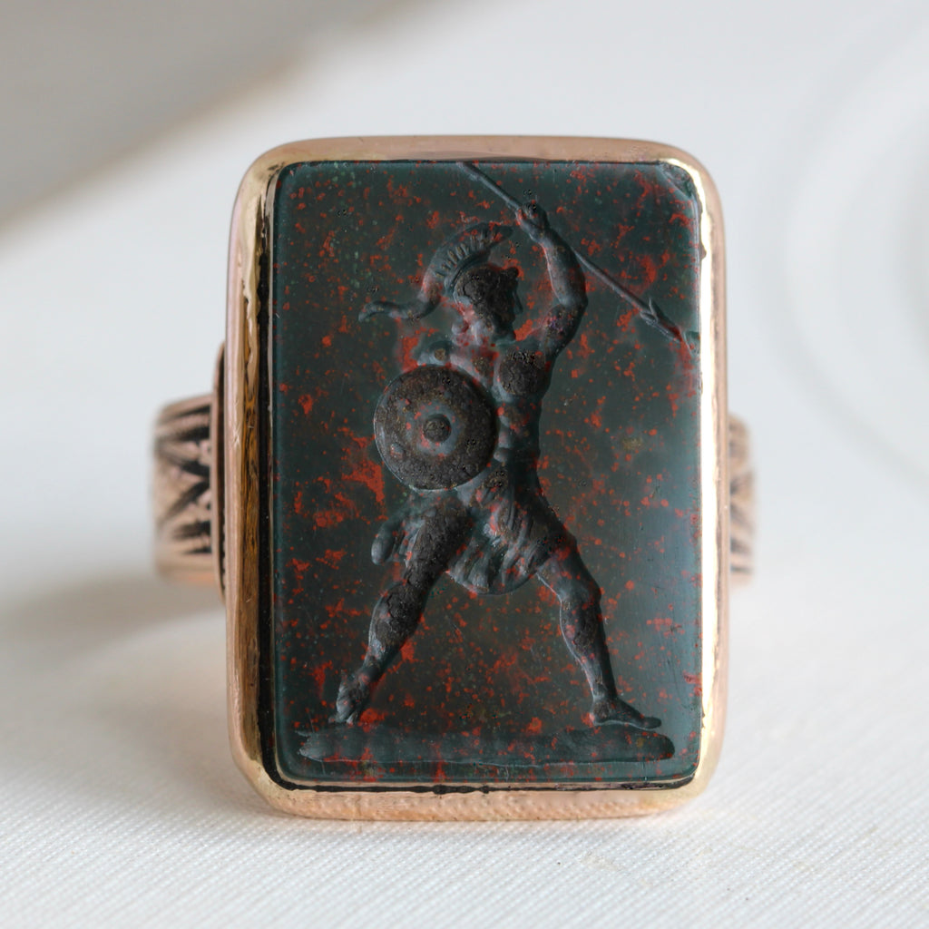 large rectanguar green stones with red spot carved with a warrior holding a spear and shield, set in a rose gold ring mounting