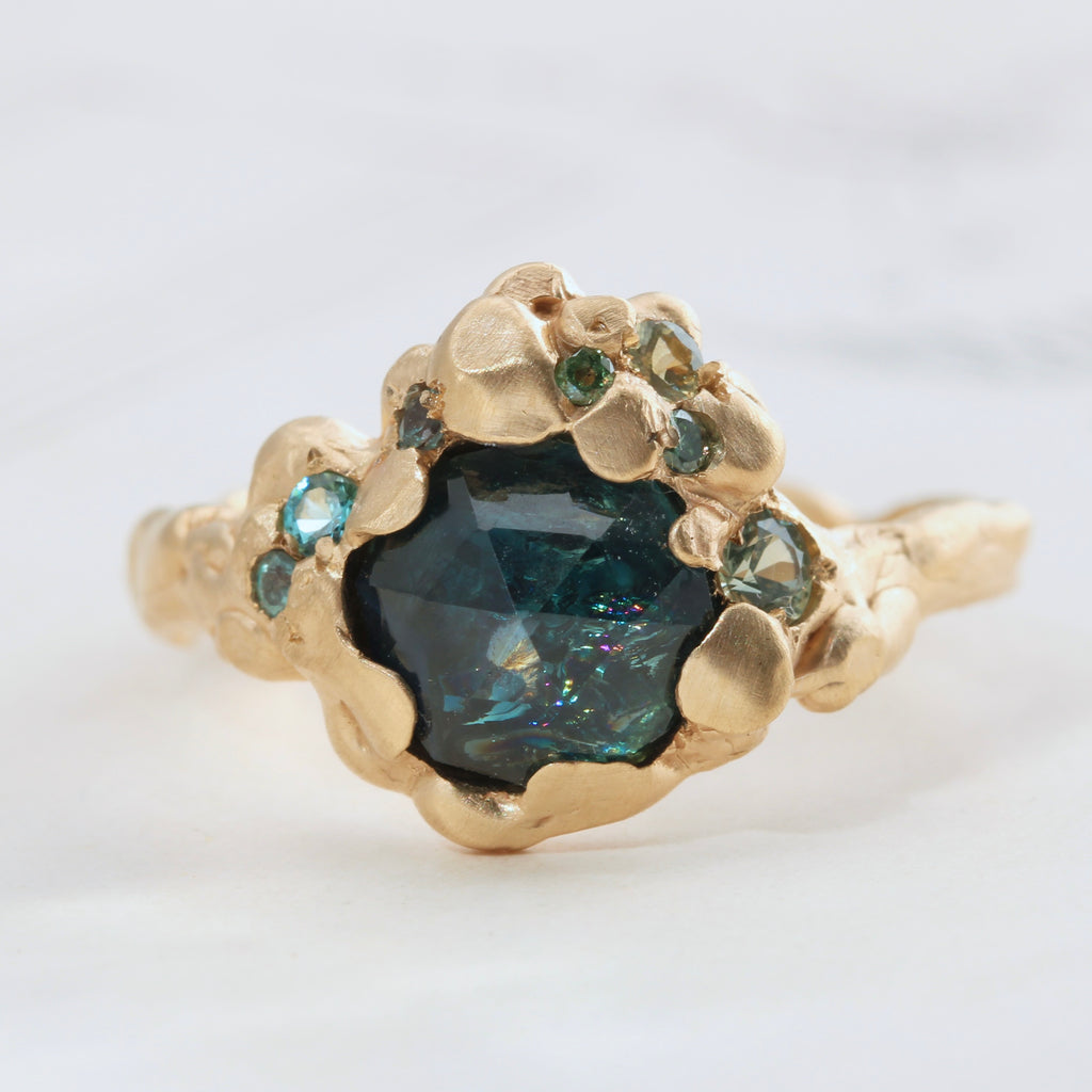 a teal green blue rose cut sapphire with rainbow colored inclusions set in an organic branch style yellow gold mounting with green gem accents