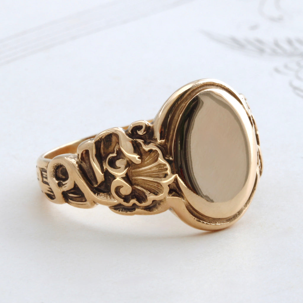 yellow gold ring with a blank oval face and a design of snakes and scallop shells carved into the shoulders