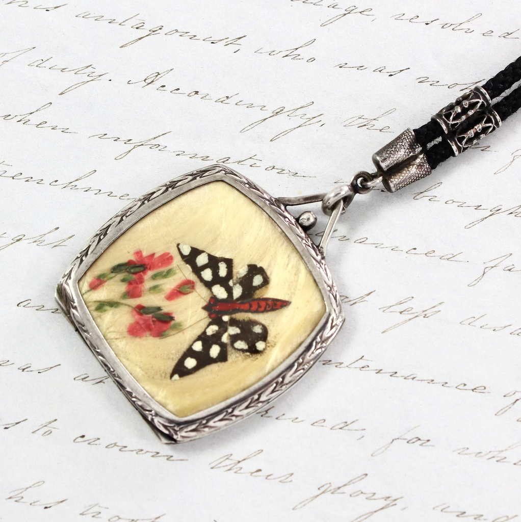 Antique sterling silver locket with one side featuring an image of a butterfly flying towards a bunch of flowers, composed of real butterfly wing pieces. Hanging on a black fabric rope necklace.