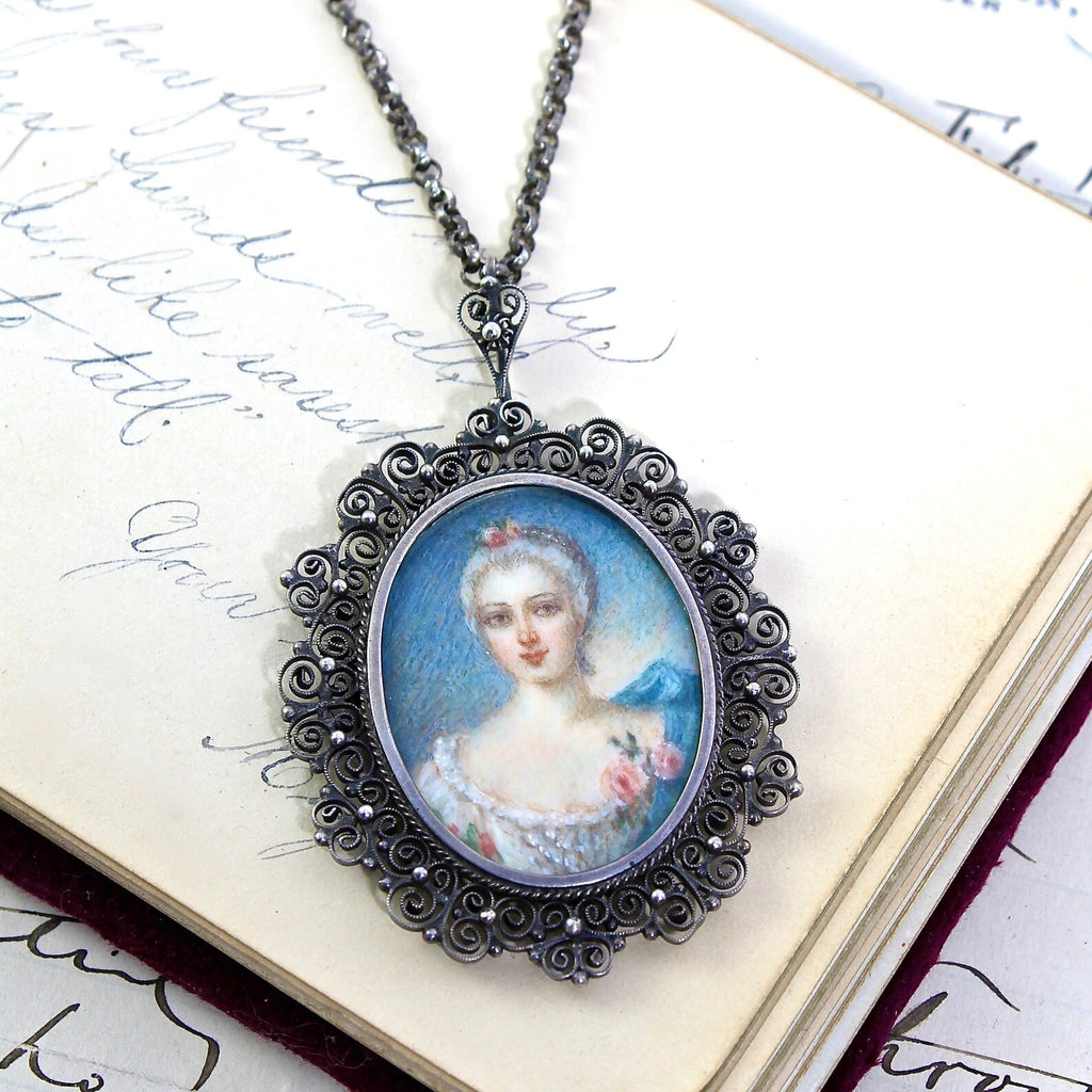 Sterling silver pendant with wirework filigree scroll design border and a hand painted image of a woman with roses and a blue background, on a sterling silver cable chain.