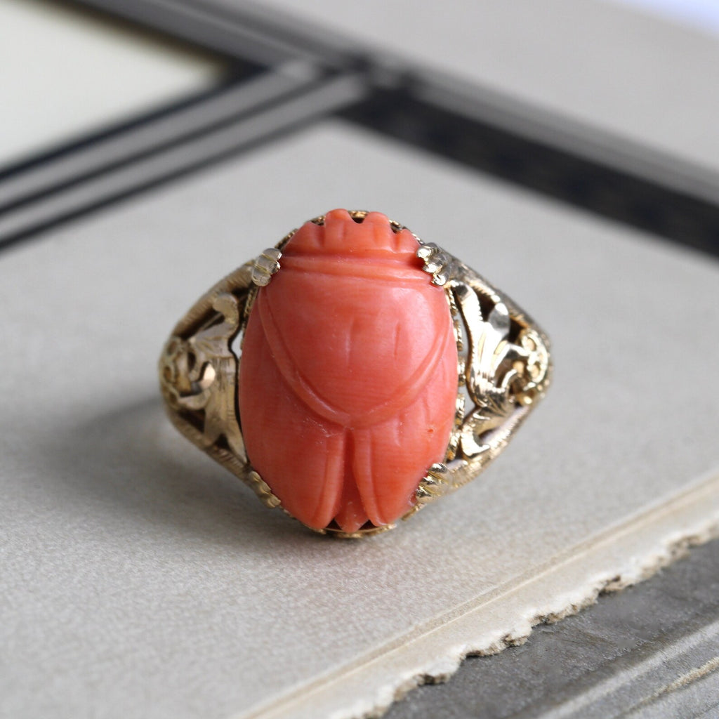 Antique yellow gold ring with a pink coral carved scarab in a prong setting with floral design cutouts.
